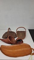 Photo showing five woven baskets including a large leaf-shaped tray with damage at the tip, curved basket with handle, square basket with handle, small semi-circular basket, and large round basket.