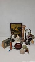 Array of decorative items displayed on white surface including candlestick, metal basket with flowers, framed painting, Christmas ornaments, ceramic and glass pieces.