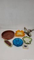 Seven decorative items arranged on flat surface including glass swan, multiple glass bowls in amber, green, and blue colors, ceramic owl figurine, small botanical dish, and terra cotta pot.