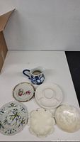 View of porcelain items including small pitcher with blue floral design and various decorative plates on table.