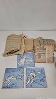 Multiple blue and white decorative tiles with hand-painted figures and nature scenes, some still wrapped in newspaper