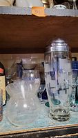 Martini shaker with etched glass pattern and metal top, clear double-bowl glass container with rounded bottom, placed on shelf with bottles and glasses nearby.