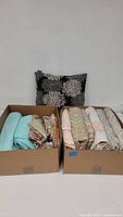 Lot showing two boxes containing multiple folded linens with various patterns and one floral decorative throw pillow behind the boxes.