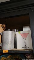 Shelf showing silver textured pitcher alongside a box featuring a pink martini glass with twisted stem design.