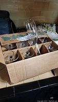 Photo showing a cardboard box with eight clear stemmed wine glasses arranged in two rows with one glass held upright to display the shape.