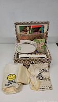 Full view of closed and open patterned sewing box with contents including fabric bags, sewing accessories, and books.