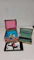 Two vintage jewelry boxes, one fabric-covered with turquoise interior holding porcelain figurines, one green with satin-lined compartments, and a silk scarf tied with a black ribbon.