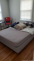 Sultan boxspring bed frame with attached feet and mattress with bed linens piled at one end of the bed next to a red office chair in a bright room with hardwood floor and windows with blinds.