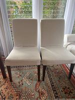 Front view of two white slipcovered dining chairs with dark wooden legs, placed on patterned rug by window.