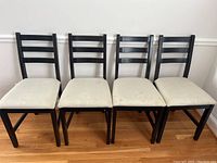 Front view of the four black wooden chairs with light stained upholstered seats, aligned side by side.