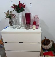 Photo of vases, candles, faux flowers arranged on a white chest including clear and colored glass vases, several candles, and two arrangements of faux flowers.