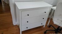 Full front view of the white dresser showing three drawers with black knob handles, standing on wooden floor against a white wall.