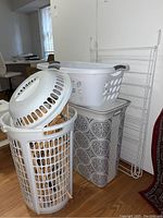 Photo of stacked laundry baskets and hampers including two large white plastic baskets and one beige patterned fabric hamper. White plastic hangers visible in the baskets.