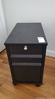 Top-side photo showing the black rectangular file cabinet with three drawers and a key lock on the top drawer, placed on a wooden floor.