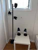 Photo of one black metal floor lamp with three adjustable lamp heads and two small black table lamps on a small white table near a window