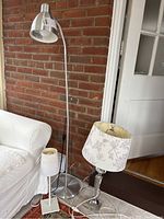 Photo showing all three lamps together: one tall chrome floor lamp with curved neck, two smaller table lamps with floral patterned shades and chrome bases near a brick wall and white couch.