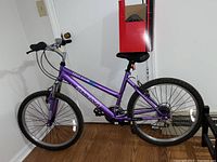 Purple Magna Great Divide mountain bike with front suspension, flat handlebars, knobby tires, and black saddle. Bike is shown against indoor wall.