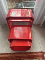 Four red nesting tables stacked nested together on tiled floor in front of glass door. Tables have rounded edges and glossy paint finish.