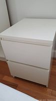 White night stand from front showing two drawers and handle design, placed on wood floor near white wall.