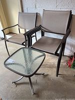 Four outdoor chairs with brown mesh fabric and dark metal frames, stacked two on two, adjacent to a small square glass-top side table with dark metal legs. Concrete floor and cinder block wall background.