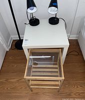 Photo showing two side tables: a white square table and a smaller wooden table with a glass top. Also visible are two black lamps on the white table.