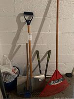 Full view of all yard tools leaning against a cinder block wall: snow shovel, leaf rake, clippers, small shovel, and bag of salt on floor