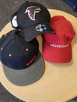 Top view of three sports caps showing front logos - black Falcons cap, red Air Jordan cap, black LeBron James cap with patterned brim