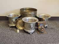 Three brass planters, large brass pot, brass plate, small brass bell, and steel piano man music box grouped on a carpeted floor.