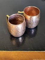 Two hammered copper mugs with rounded bodies and brass handles, showing natural tarnish and patina, sitting on a textured dark surface.