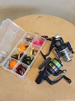 Clear plastic tackle box open showing nine compartments with assorted soft plastic fishing lures, jig heads, hooks and bobbers, next to two fishing reels.