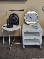 View of all items together showing the round steel table, steel rolling cart, black desk lamp, black Honeywell tabletop fan, white wall clock, and metal pencil holder on the cart.
