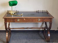Front view of wood office desk with green banker lamp placed on top, showing curved legs and black top surface with some scratches.