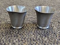 Two pewter shot glasses shown side by side, viewed from the front on a carpeted floor. Both have flared tops and rounded bases with some distortion. Visible texture and metallic finish.