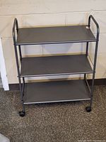 Full view of the 3-tier rolling metal storage cart against a wall with four casters visible.