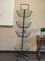 Full view of the black metal tiered wire basket storage unit on casters with four stacked baskets and sign holder on top, against a wall.