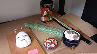 Photo showing white lacquered face mask, orange ceramic mug with broken handle, cloisonné container with lid, hand-painted small container with lid, chopsticks with decorative case