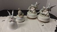 Set of 3 ceramic bird music carousels and 1 ceramic bell arranged on a table with a white cloth, highlighting their design and size.