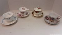 Four fine china cups and saucers arranged for display. Each cup is paired with its matching saucer. Floral designs with gold trim are visible on each set.
