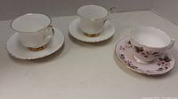 Three cups and saucers on a flat surface with two white Royal Albert Val Dor cups and saucers and one pink floral Colclough cup and saucer.