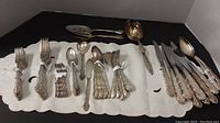 Photo showing multiple flatware pieces including forks, spoons, and knives arranged neatly on a white cloth, highlighting the ornate handles and polished condition.