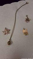 Four gold jewelry pieces arranged on fabric: chain necklace with oval locket, filigree leaf pendant, solid maple leaf brooch, square religious pendant