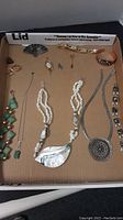 Wide shot of assorted jewelry including necklace with abalone pendant, silver necklace with ornate pendant, green stone bracelet and earrings, pink carved ring, and other rings and pins.