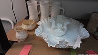 Wide view showing large fluted hobnail milk glass bowl, three tall milk glass mugs, and Belleek cream and sugar on cardboard.