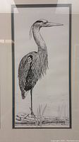 Full view of framed heron ink drawing showing bird standing in a wetland area with reeds and rocks.
