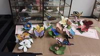 Photo showing all porcelain butterfly figurines arranged on the table, each mounted on colorful flower bases, with some butterflies in vibrant colors including yellow, purple, and blue.