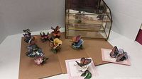 Nine porcelain butterfly figurines displayed on cardboard with flowers and realistic painted details, various colors including orange, purple, blue, and yellow.
