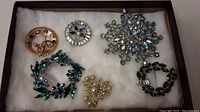 Six assorted rhinestone brooches on white fabric background showing variety of shapes, colors, and materials.