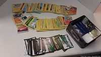 Overview of approximately 200 Pokémon trading cards spread on table with partially visible original tin box containing the cards.