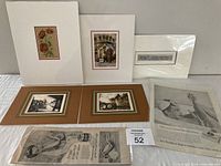Multiple framed and matted art pieces and vintage newspaper advertisements displayed laid out on a white surface