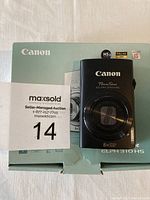 Canon PowerShot ELPH 310 HS camera shown on top of original box with auction label visible